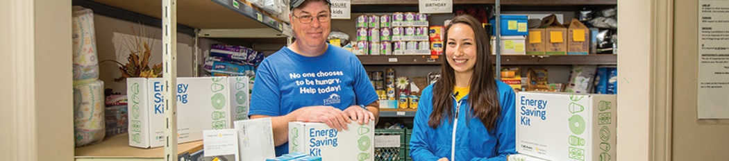 A FortisBC street team member and Food Bank volunteer smiling and holding free Energy Savings Kit boxes in a food bank