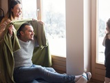 Parents and their little daughter playing peek-a-boo with a blanket by a window, snowy winter scape outside. (20-171.3 12/2020)