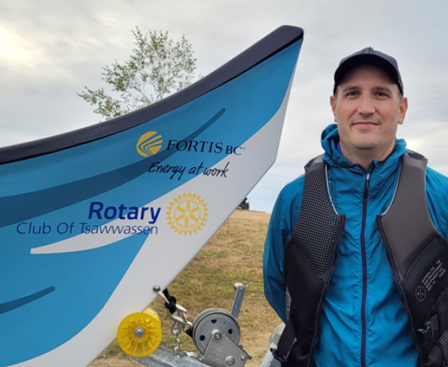 A man in a life vest and rain gear standing next to a blue and white Coast Salish canoe. 