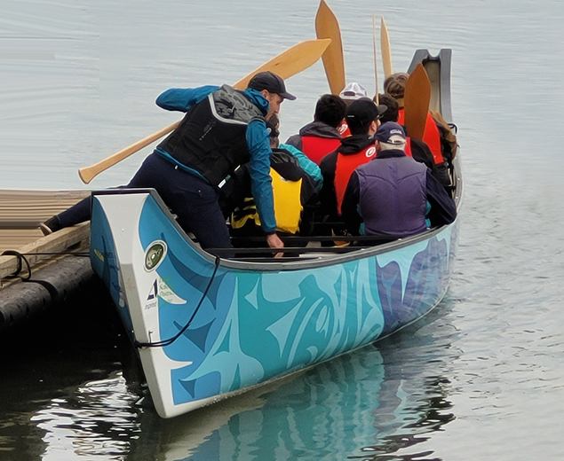 A man joining other paddlers aboard a canoe with blue and white Coast Salish artwork.
