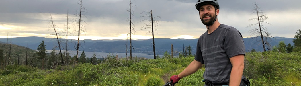 Todd Brunner standing with his bike on a trail with a lake and mountain in the background