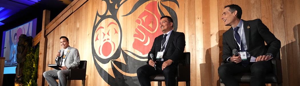 Chief Ian Campbell and FortisBC CEO Roger Dall’Antonia smiling and engaging in conversation on an event stage with an wooden backdrop showing indigenous art.