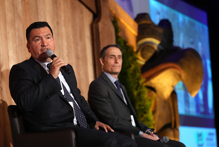 Chief Ian Campbell and FortisBC CEO Roger Dall’Antonia smiling and engaging in conversation on an event stage with an wooden backdrop showing indigenous art.