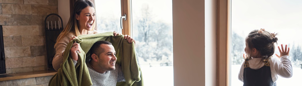 A dad and mom are playing peek-a-boo with their daughter using a blanket; the weather outside is snowy and cold.