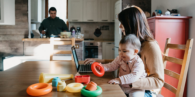 A mom is entertaining her baby on her lap with some ring toys while reading on a laptop. Her husband is in the background doing dishes.