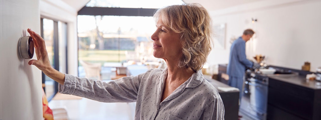 Woman reaching to adjust her thermostat while a man cooks in the background.