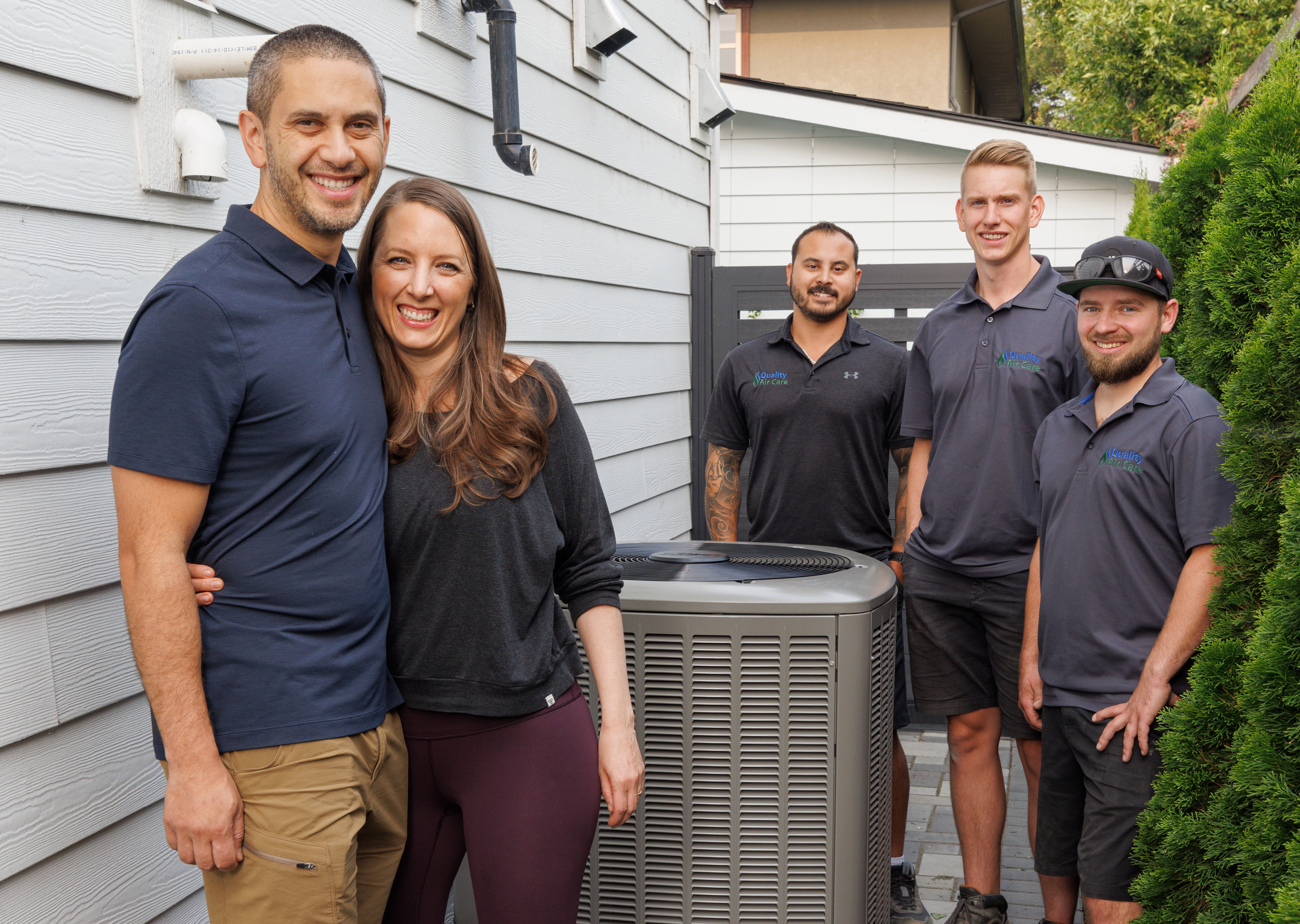 A couple standing proudly in front of their newly installed heat pump. Three contractors stand in the background