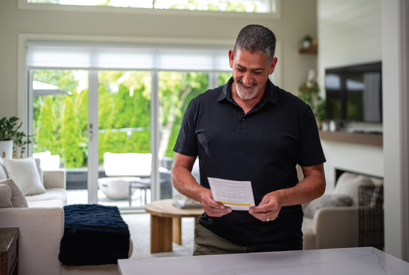 A man stands in his living room, reading an information sheet. He is smiling.