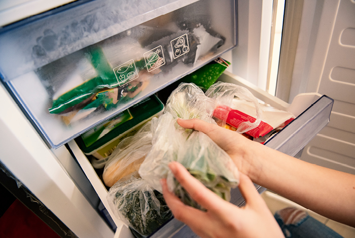 A person’s hand holding a vegetable with the refrigerator door open, showing more vegetables in the fridge drawer.