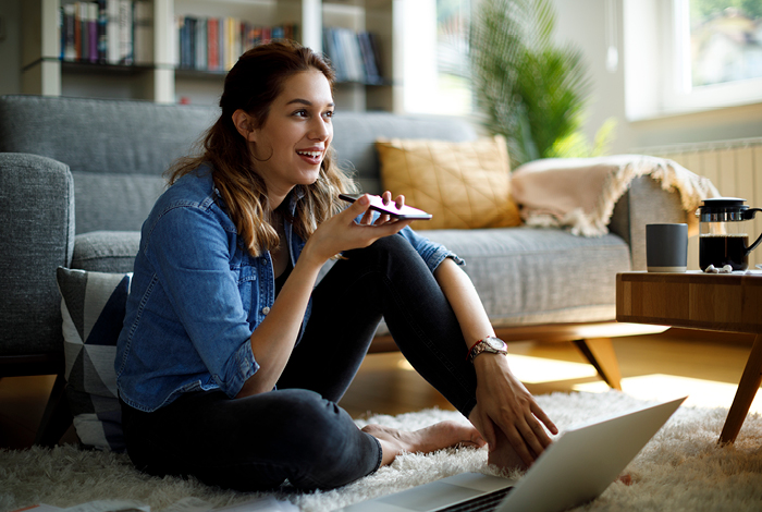 Person sitting on the floor in a cozy living room, using a smartphone and laptop, with a sofa and bookshelves in the background.