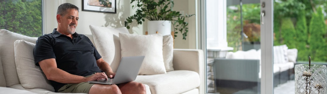 A man in a black shirt using a laptop while sitting on a white couch next to a sliding door