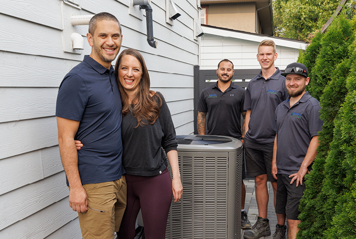 26-144-5-dualfuel-relaunch-blog-inline-700x470-7 A couple standing proudly in front of their newly installed heat pump. Three contractors stand in the background.