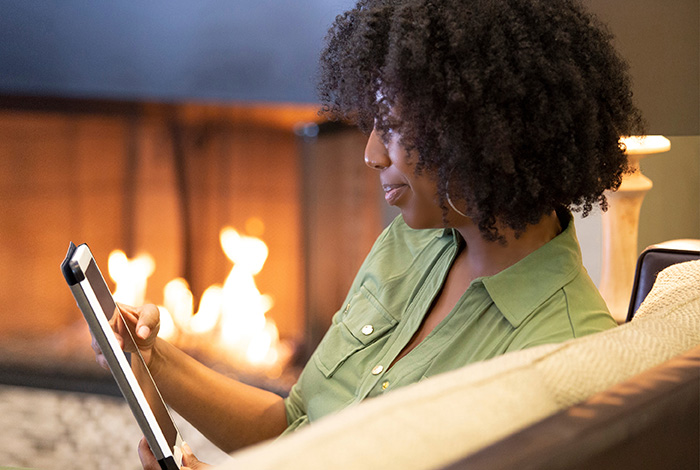 A woman looking at her tablet while sitting on a sofa with a cosy fireplace in the background.