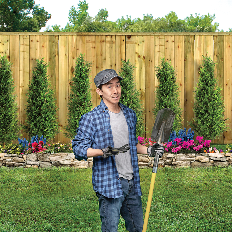 A person standing in a backyard, shrugging his shoulders and holding a shovel. Background features a wooden fence, neatly trimmed green shrubs, and colorful flower beds with pink, blue, and red blossoms.