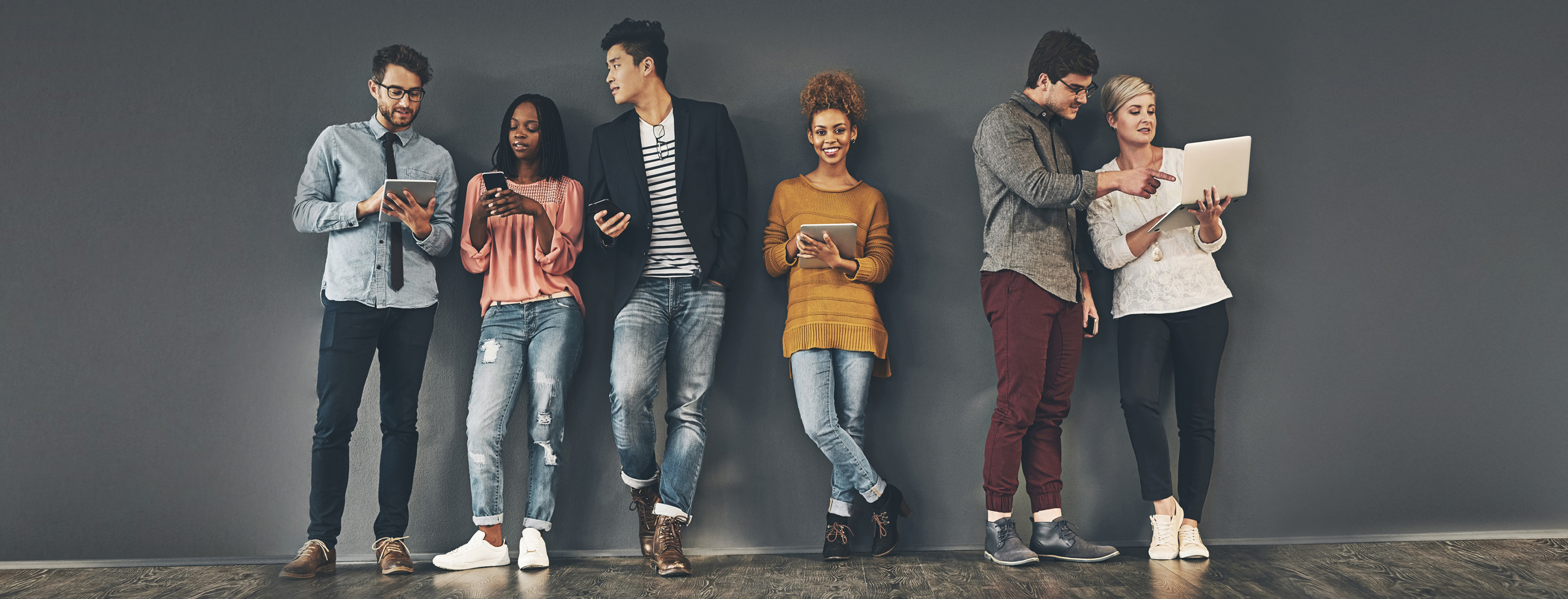 A group of people leaning against a dark grey wall, sharing information (18-150.11)