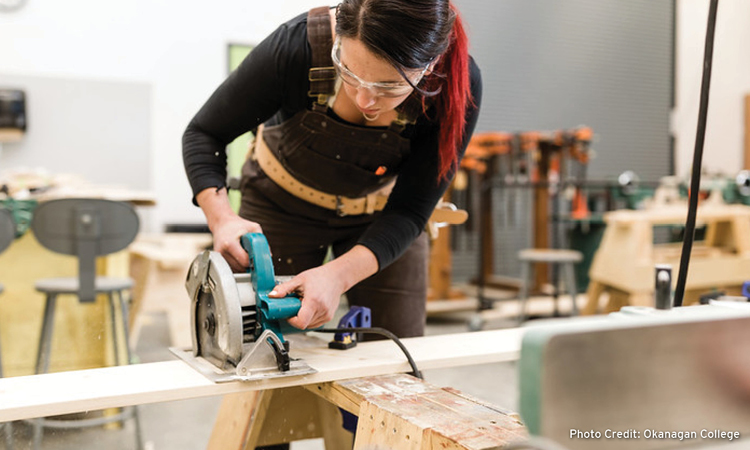 An Okanagan College student working in the wood workshop, cutting a wooden plank with a circular saw.